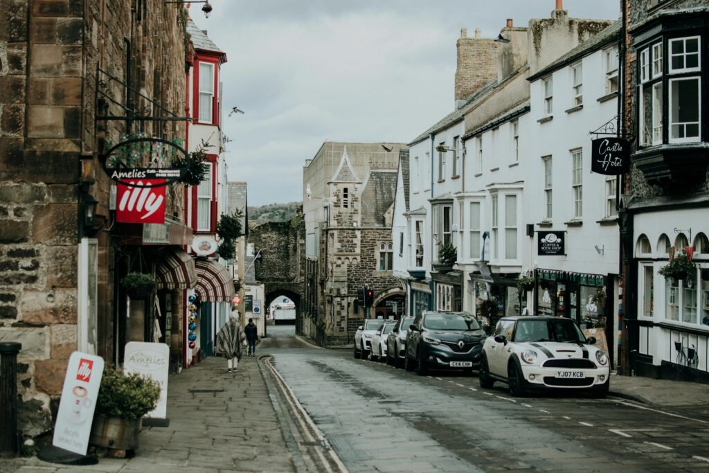 A quaint British street lined with shops and parked cars leading to an old stone gate.