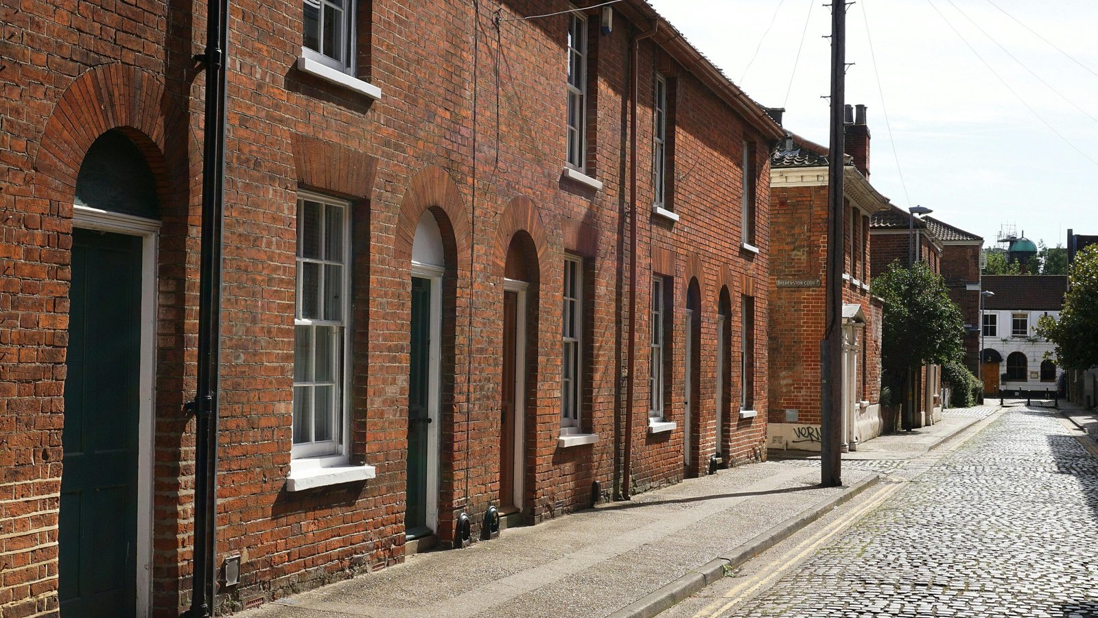 Sunlit view of traditional brick homes lining a cobblestone street in Norwich, UK.