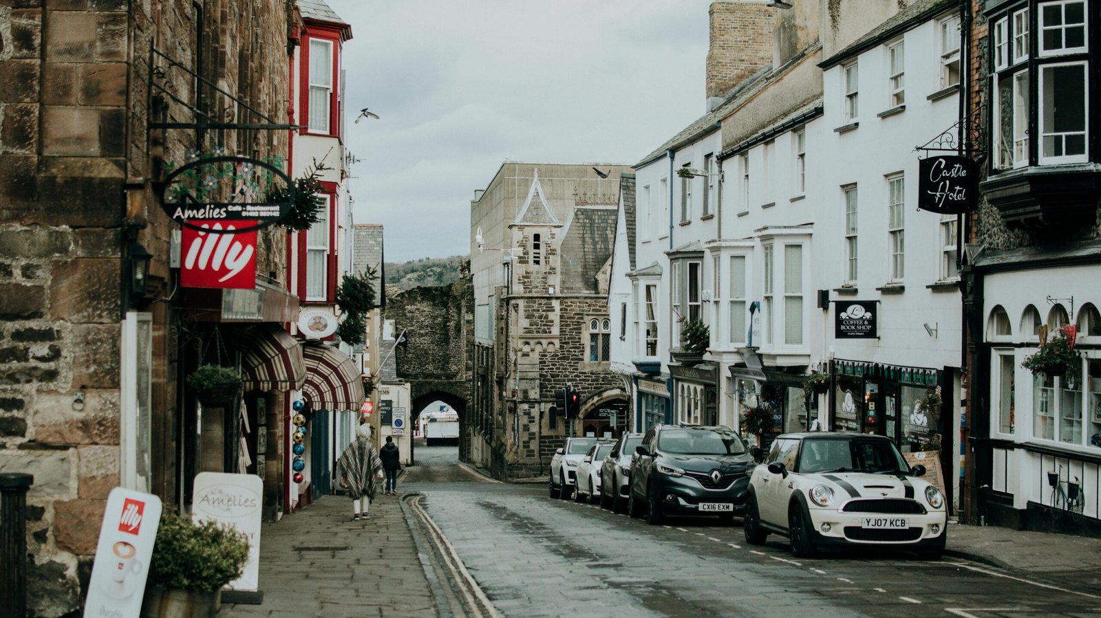 A quaint British street lined with shops and parked cars leading to an old stone gate.